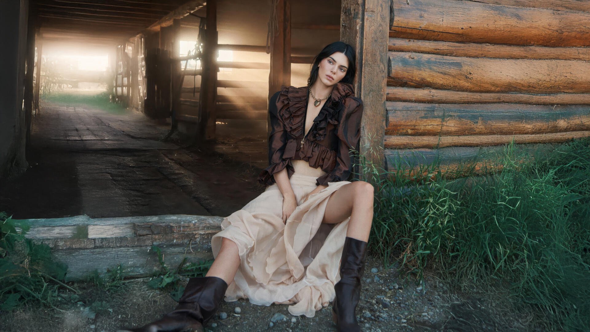 Seated female model in a flowing beige skirt and boots, leaning against a log cabin wall with sunlit barn corridor behind her — 5K Ultra HD PC desktop wallpaper.