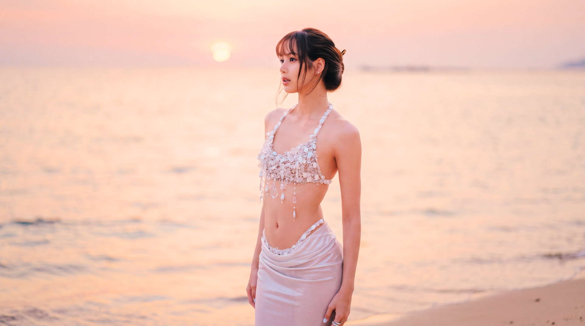 5K Ultra HD desktop wallpaper: a K-pop idol standing on a sunset beach in a white beaded outfit, pastel sky and calm sea bathed in warm golden light.