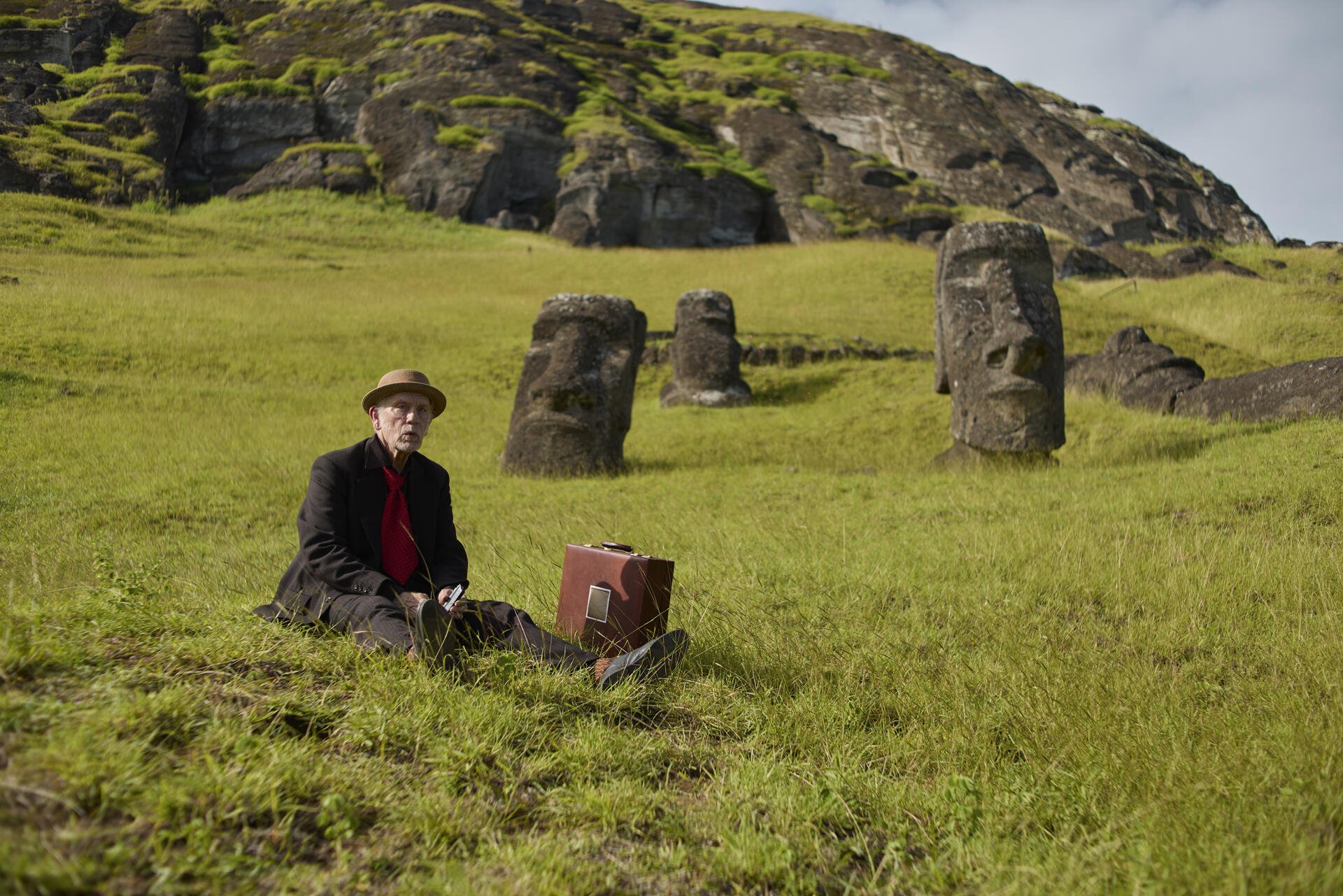 2K Quad HD PC wallpaper Wild Horse Nine: a man in a hat sits with a suitcase among moai-like stone statues scattered on a windswept green grassland.