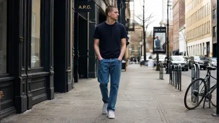 Man in normcore outfit—black tee, blue jeans and white sneakers—walking down a city sidewalk; 4K Ultra HD PC desktop wallpaper/background.