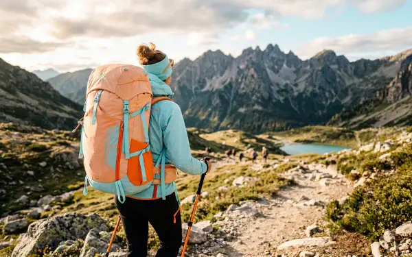 Backpacking hiker with orange pack on a mountain trail toward jagged peaks and alpine lakes — 5K Ultra HD PC desktop wallpaper and background.