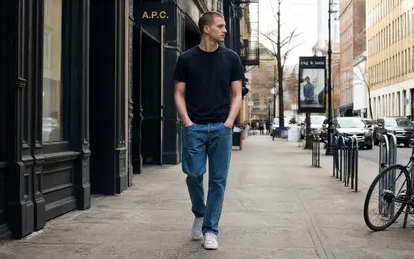 Man in normcore outfit—black tee, blue jeans and white sneakers—walking down a city sidewalk; 4K Ultra HD PC desktop wallpaper/background.