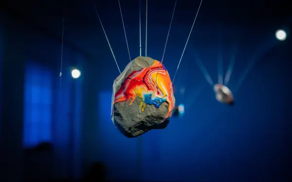 Rock painting: vivid red, yellow and blue swirls on a stone suspended by fine wires against a deep blue bokeh — 5K Ultra HD PC desktop wallpaper and background.