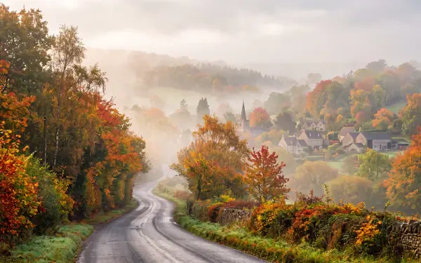 Foggy morning country road winding through autumn trees to a mist-shrouded village — 5K Ultra HD PC desktop wallpaper and background.