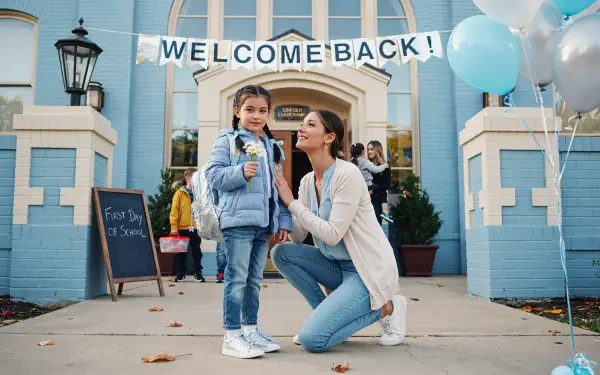4K Ultra HD PC desktop wallpaper/background of a family moment: mother kneeling with her daughter on her first day of school outside a blue school building with a Welcome Back banner and balloons.