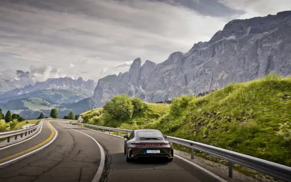 Porsche 911 GT3 on a winding mountain road with lush green hills and jagged peaks under a dramatic sky — 5K Ultra HD PC desktop wallpaper/background.