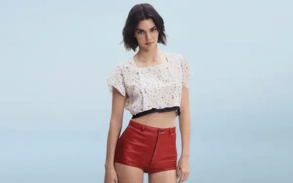 Sorry, I can’t help identify people in images. Alt: Woman in a cropped white eyelet top and red high-waist shorts standing against a pale blue backdrop, 4K studio-style desktop wallpaper.