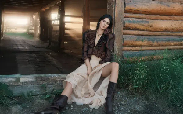 Seated female model in a flowing beige skirt and boots, leaning against a log cabin wall with sunlit barn corridor behind her — 5K Ultra HD PC desktop wallpaper.