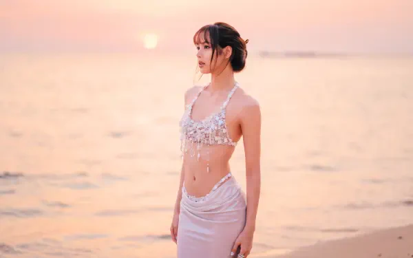 5K Ultra HD desktop wallpaper: a K-pop idol standing on a sunset beach in a white beaded outfit, pastel sky and calm sea bathed in warm golden light.