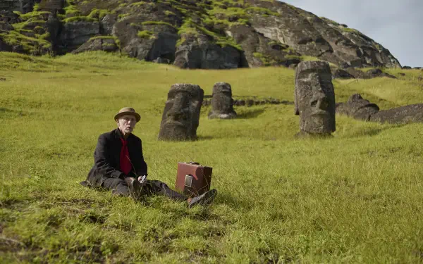 2K Quad HD PC wallpaper Wild Horse Nine: a man in a hat sits with a suitcase among moai-like stone statues scattered on a windswept green grassland.