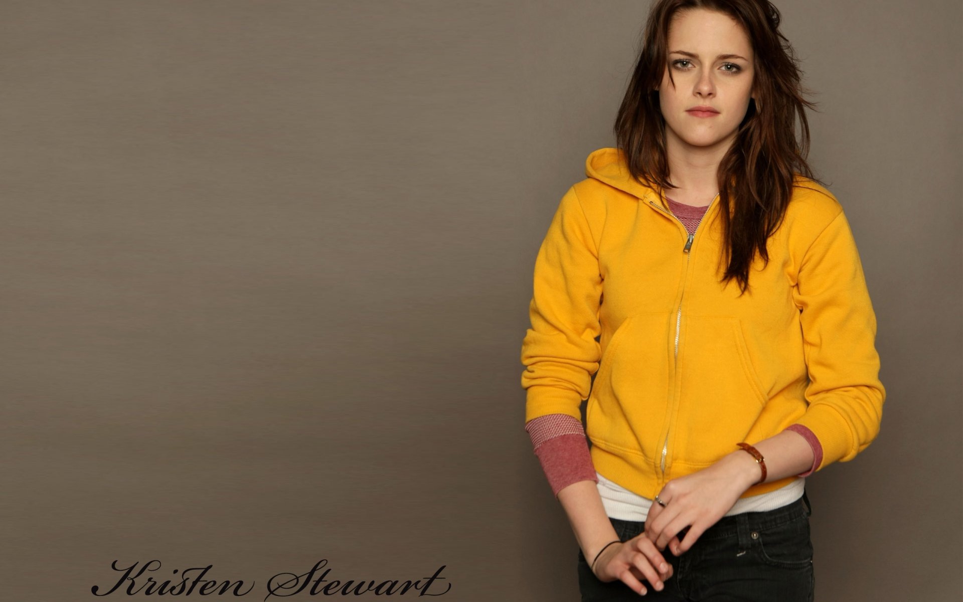 A high-definition wallpaper featuring Kristen Stewart, wearing a bright yellow hoodie, standing confidently against a neutral background. Her captivating presence is highlighted in this image.
