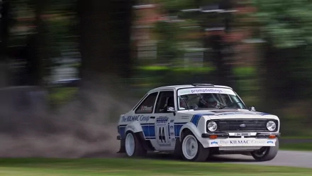 HD desktop wallpaper featuring a classic Ford Escort rally car speeding on a dirt track with motion blur and greenery in the background.
