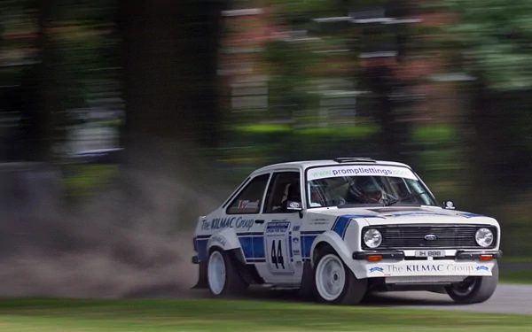 HD desktop wallpaper featuring a classic Ford Escort rally car speeding on a dirt track with motion blur and greenery in the background.