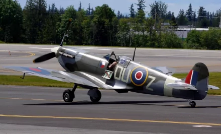 A Supermarine Spitfire aircraft taxies on the runway, set against a backdrop of lush trees and a clear blue sky, making for a striking HD desktop wallpaper.