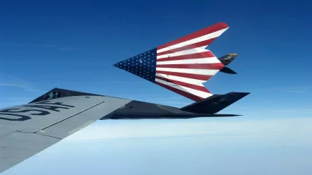 A striking HD desktop wallpaper featuring a stealth aircraft soaring through a clear blue sky, adorned with a bold American flag design, showcasing military power and innovation.