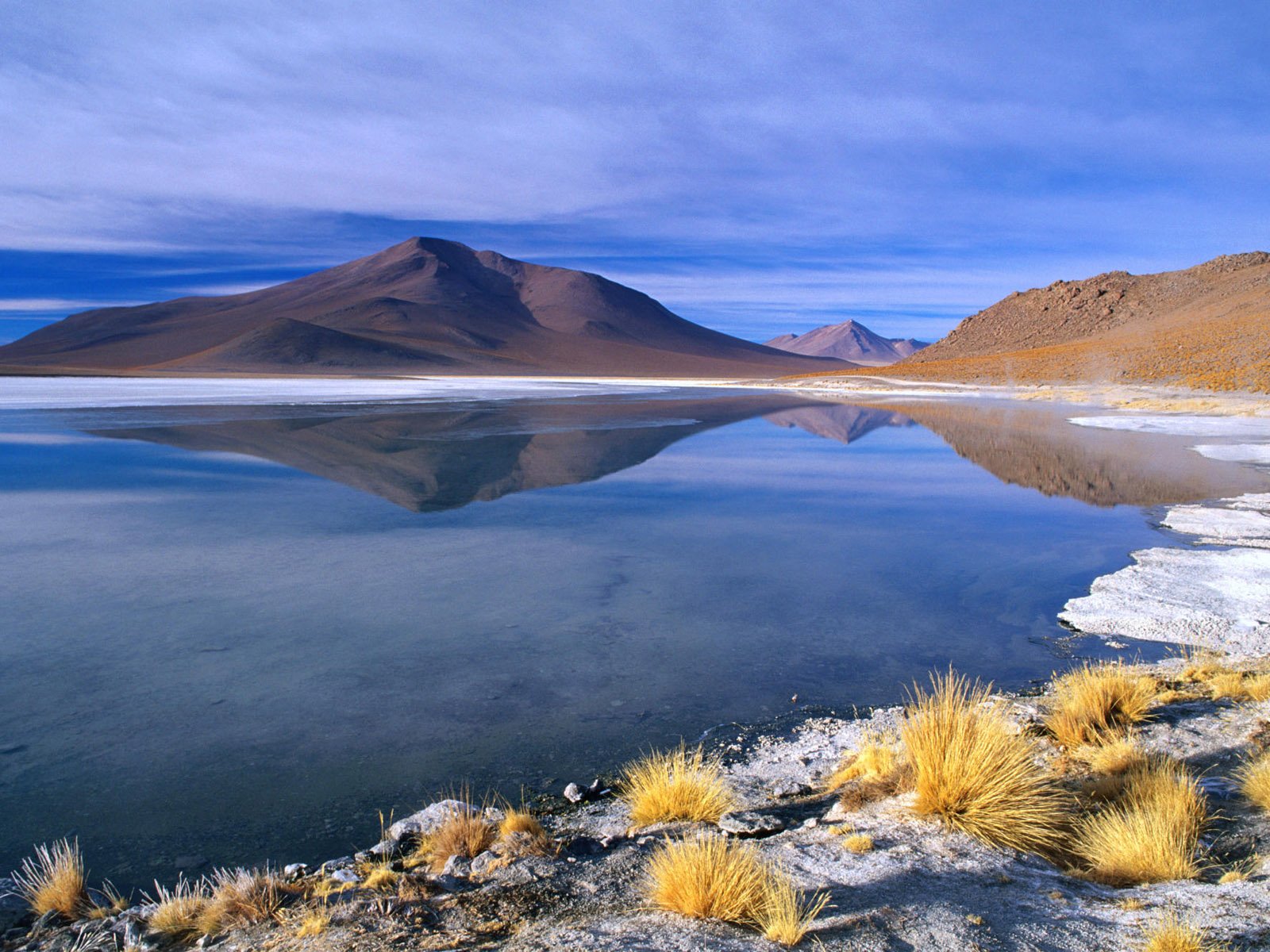 A serene lake surrounded by mountains, with a sandy beach and golden grasses, reflecting the blue sky. This HD wallpaper captures the beauty of nature in a tranquil setting.