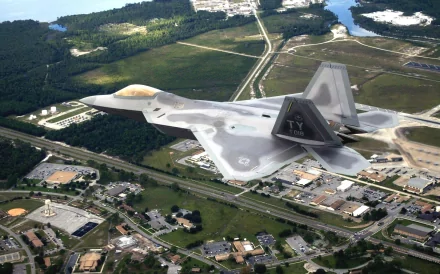 A Lockheed Martin F-22 Raptor flies over a landscape, showcasing its sleek design and military prowess, set against a backdrop of green fields and a winding road.
