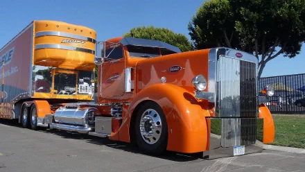 HD desktop wallpaper featuring a bright orange Peterbilt truck parked on a street with trees and clear blue sky in the background.