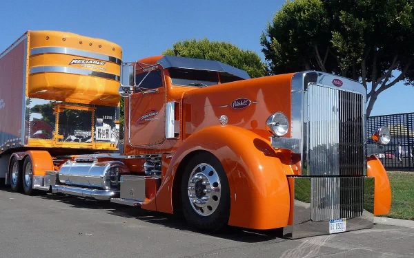 HD desktop wallpaper featuring a bright orange Peterbilt truck parked on a street with trees and clear blue sky in the background.