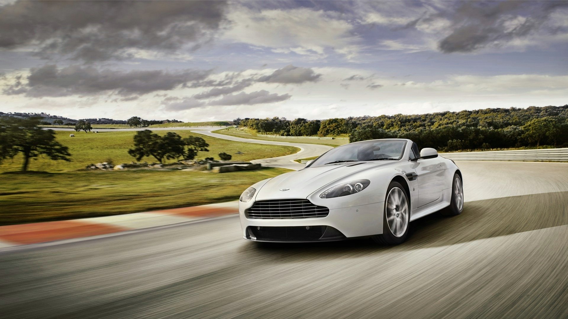 HD PC desktop wallpaper featuring a sleek white Aston Martin V8 Vantage driving on a winding road under a cloudy sky.