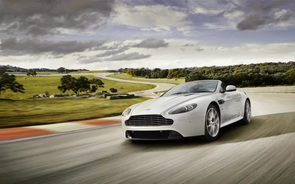 HD PC desktop wallpaper featuring a sleek white Aston Martin V8 Vantage driving on a winding road under a cloudy sky.