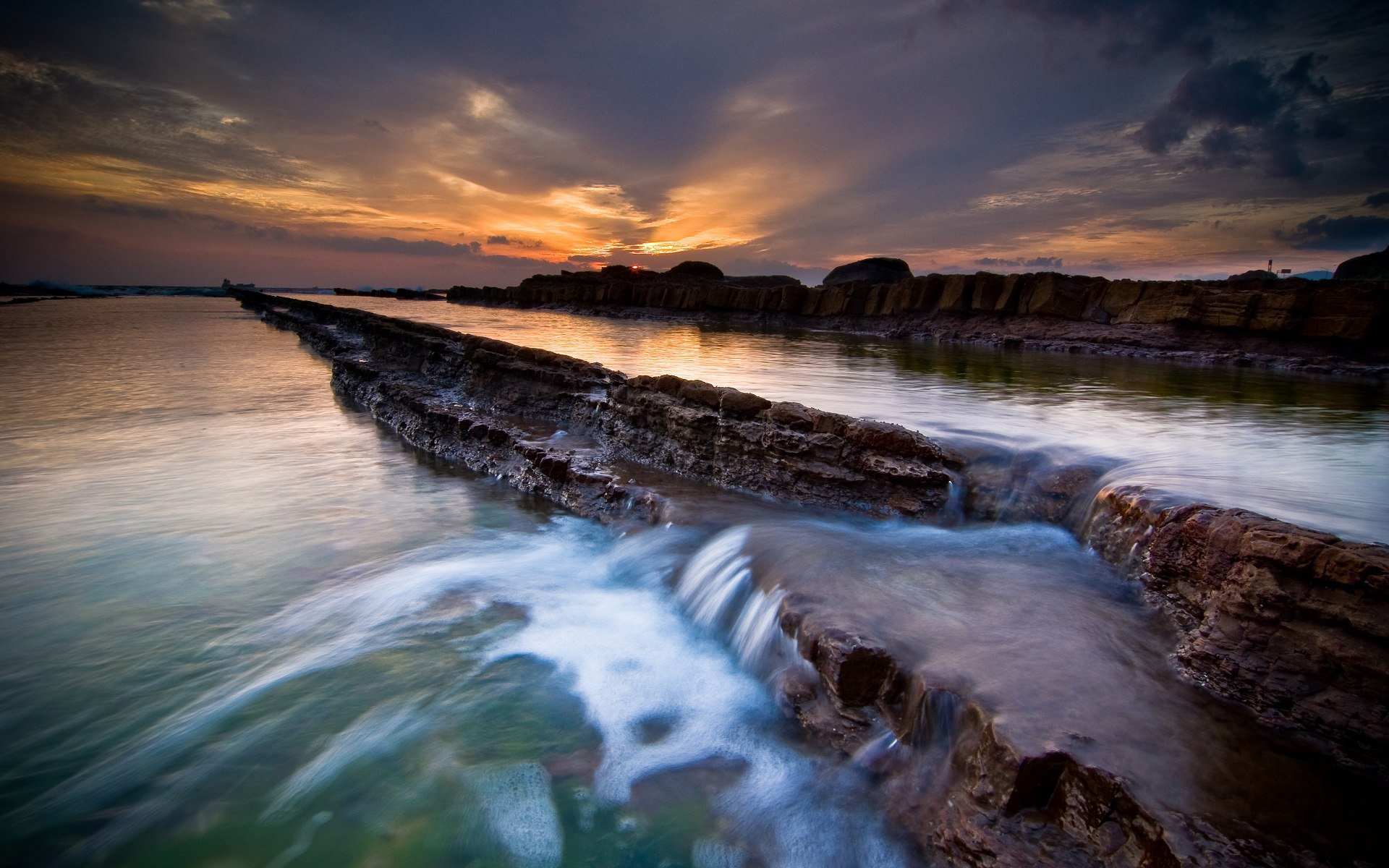 A stunning sunset illuminates a scenic waterfall cascading over rugged rocks, reflecting vibrant colors on tranquil waters, creating a captivating nature backdrop.