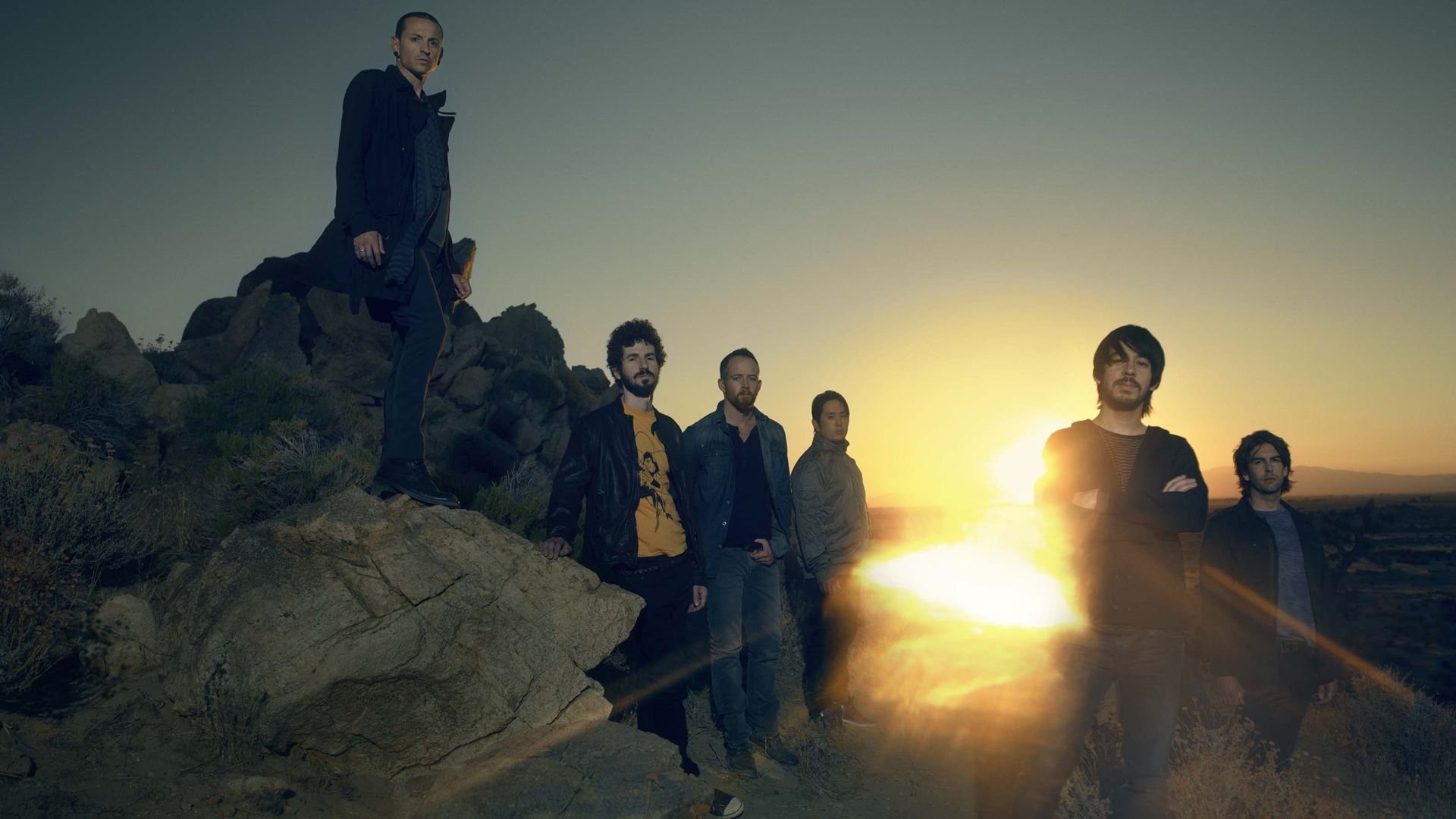 HD wallpaper featuring the band Linkin Park standing on rocks against a vibrant sunset backdrop, capturing a moment of musical artistry and energy.