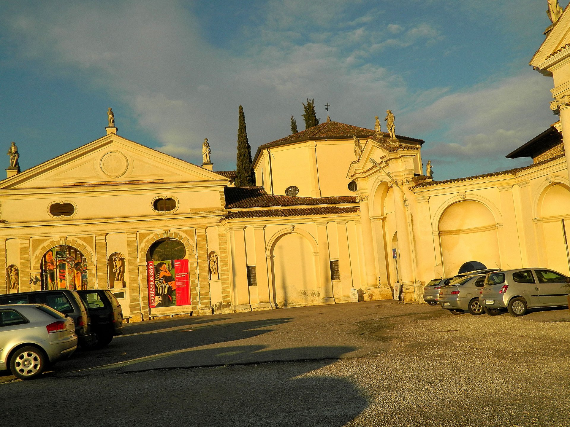 Photography of a sunlit classical plaza building with arches and parked cars under a blue sky, a 4K Ultra HD PC Desktop Wallpaper and Background image of a place.