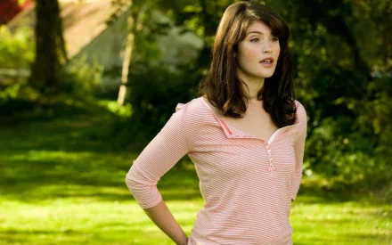 Gemma Arterton stands outdoors in a serene garden, wearing a casual pink striped shirt, embodying a relaxed and natural beauty. High-definition desktop wallpaper.