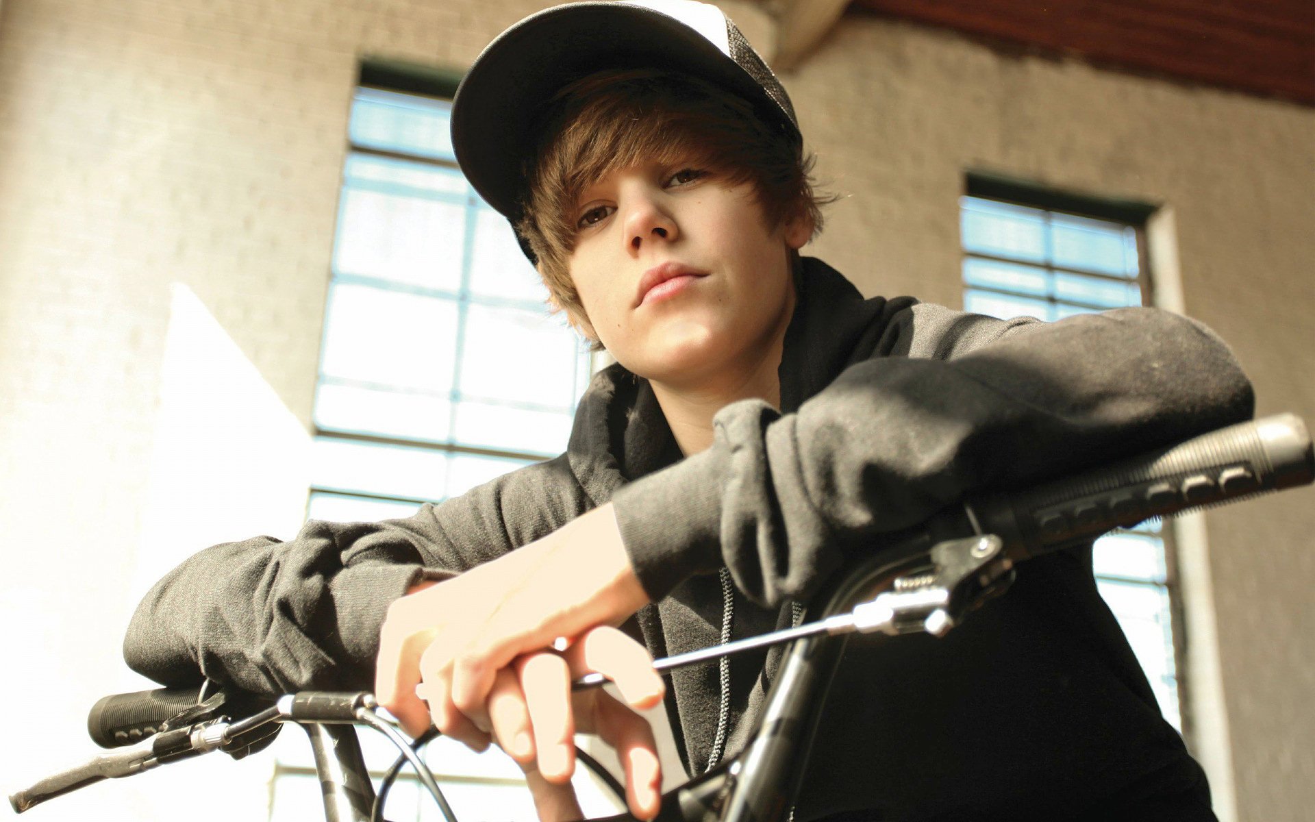 HD PC desktop wallpaper featuring a young Justin Bieber leaning on a bicycle indoors, framed by sunlit windows, capturing a casual music vibe.