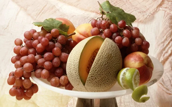 A vibrant HD desktop wallpaper featuring a fresh assortment of cantaloupe melon, red grapes, and other fruits arranged on a white plate.