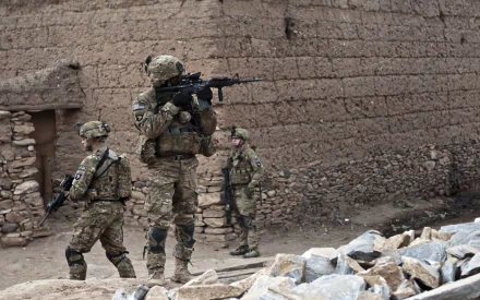 U.S. Army Infantry soldiers in full combat gear patrol a rugged war zone with mud-brick walls, captured in an HD military desktop wallpaper background.