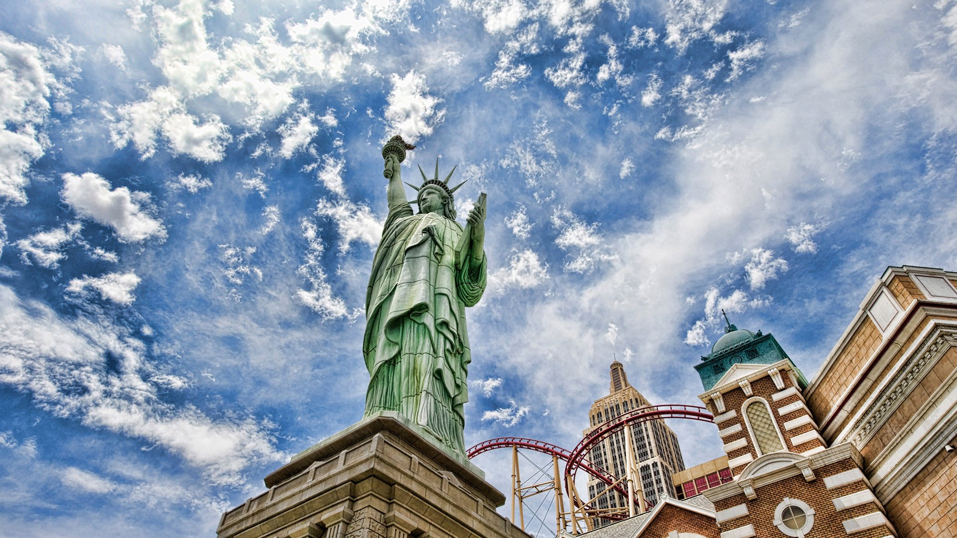 HD PC desktop wallpaper featuring the man-made Statue of Liberty against a vibrant blue sky with scattered clouds and nearby architectural structures.