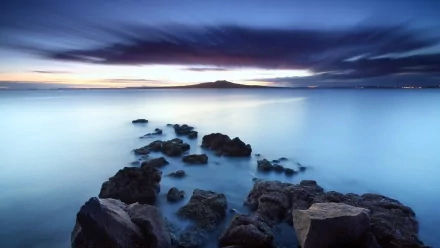 HD PC desktop wallpaper showcasing a serene seascape with calm blue waters, rocky shoreline, and a distant island under a dramatic twilight sky.