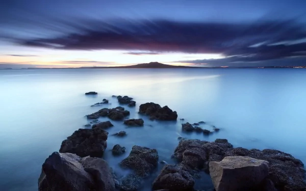 HD PC desktop wallpaper showcasing a serene seascape with calm blue waters, rocky shoreline, and a distant island under a dramatic twilight sky.