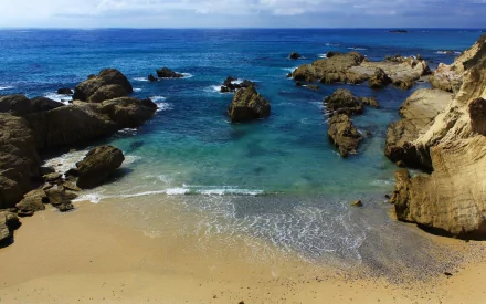 A serene seascape featuring gentle waves lapping at a sandy beach, surrounded by rugged rocks and clear blue water, offering an inviting view of nature's beauty. HD wallpaper.