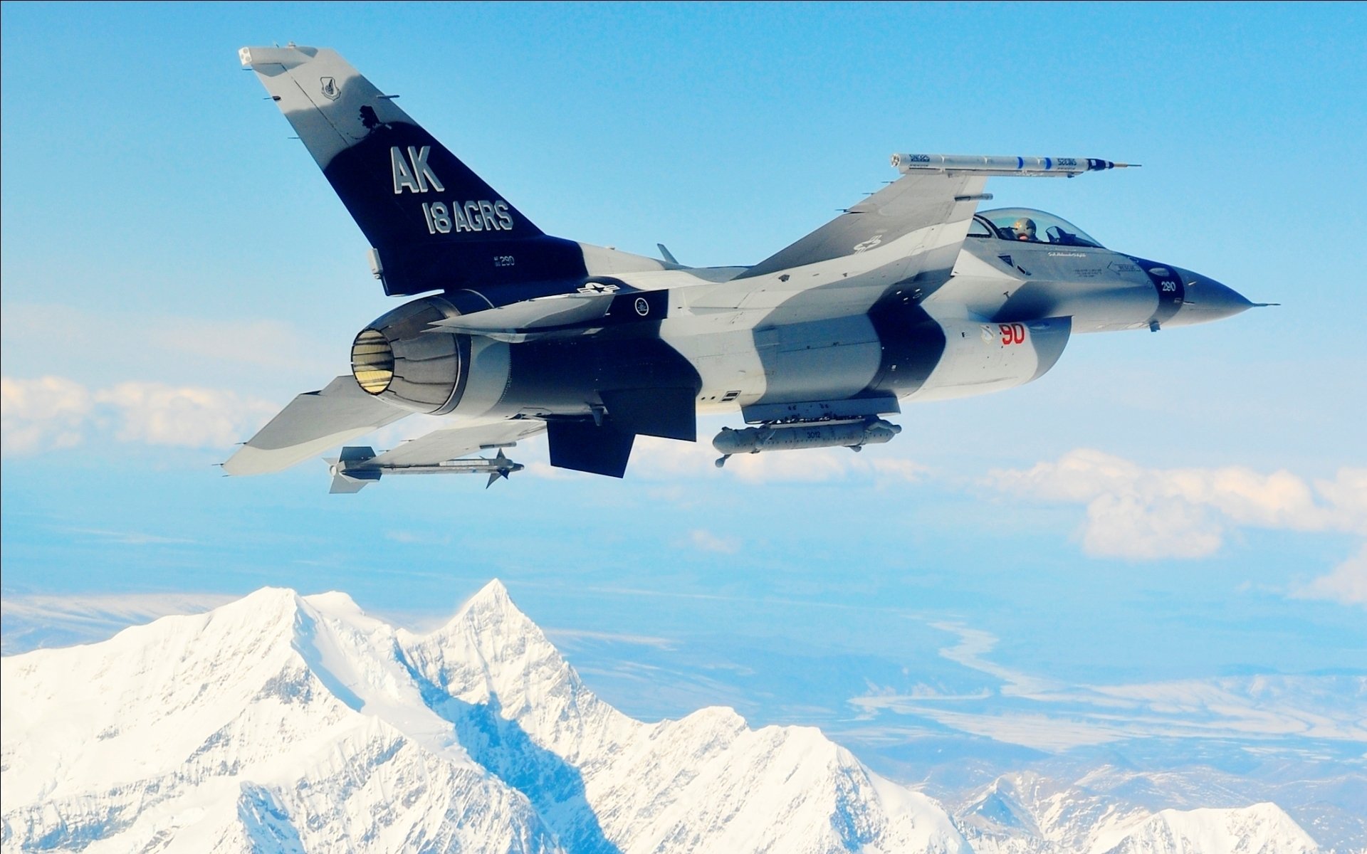 HD desktop wallpaper of a General Dynamics F-16 Fighting Falcon jet flying over snowy mountain peaks against a clear blue sky, showcasing military aviation power.