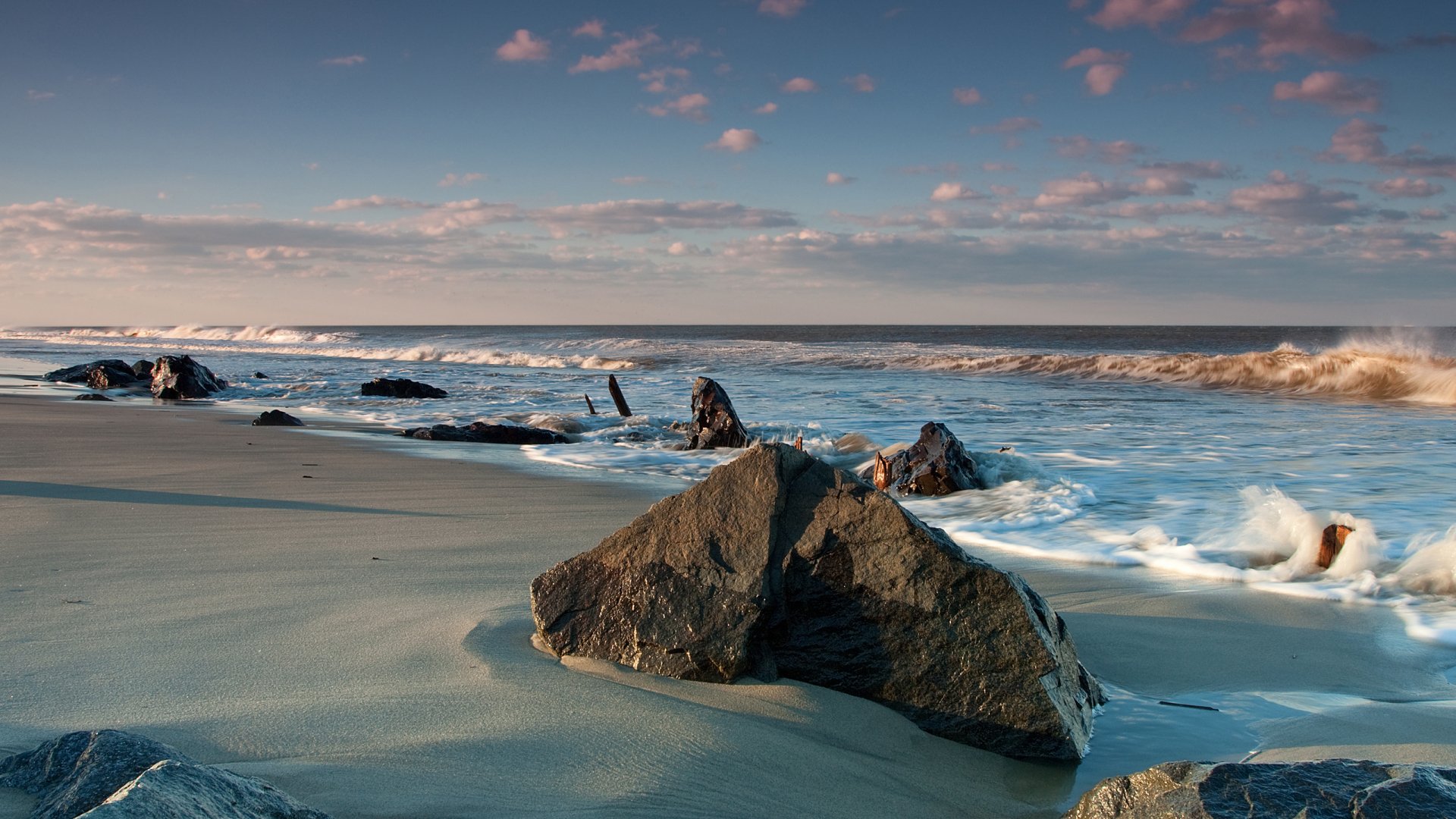 HD PC desktop wallpaper featuring a serene seascape with smooth waves, scattered rocks on the sandy shore, and a soft, colorful sky at sunset.