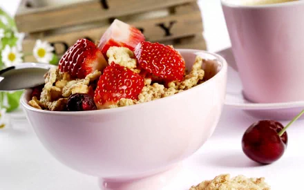 HD desktop wallpaper featuring a close-up of a breakfast bowl filled with muesli, cherry, and sliced strawberries, alongside a pink cup and fresh cherry on a white surface.