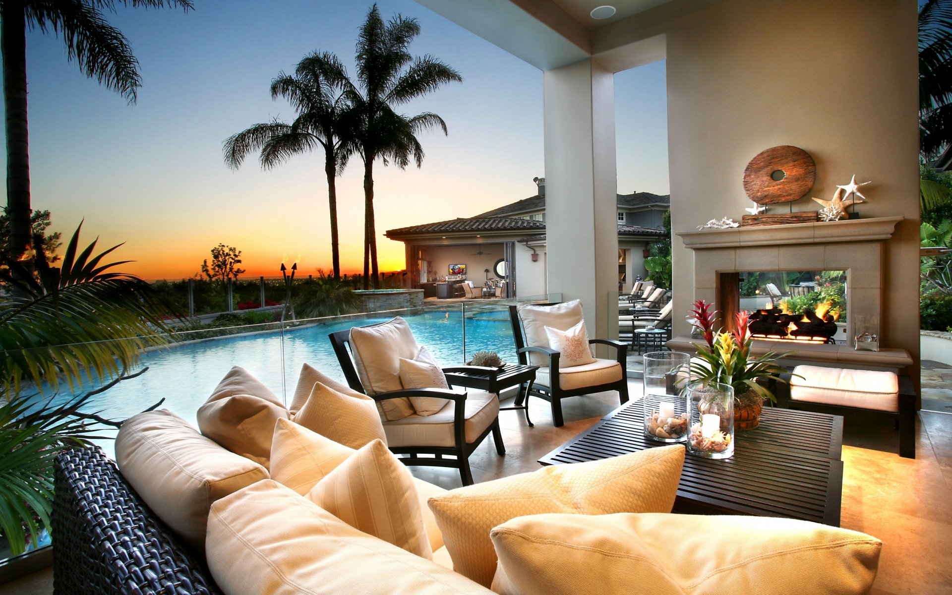 HD PC desktop wallpaper of a cozy outdoor lounge area with plush seating, a fireplace, and a pool overlooking palm trees at sunset, evoking a relaxing holiday vibe.