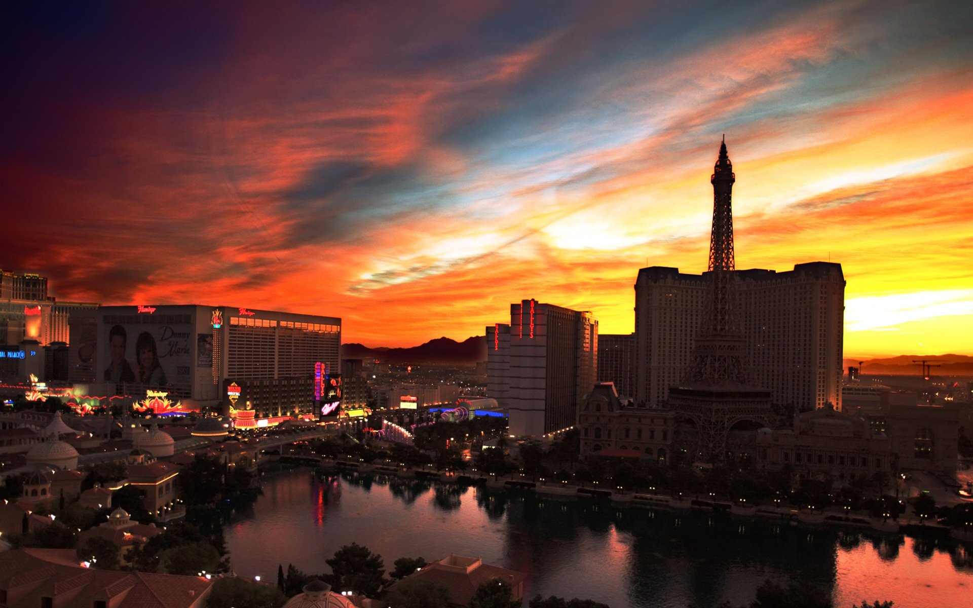HD PC desktop wallpaper and background: man-made Las Vegas skyline at sunset, neon-lit Strip reflected in waters, silhouetted Eiffel Tower replica beneath a blazing colorful sky.