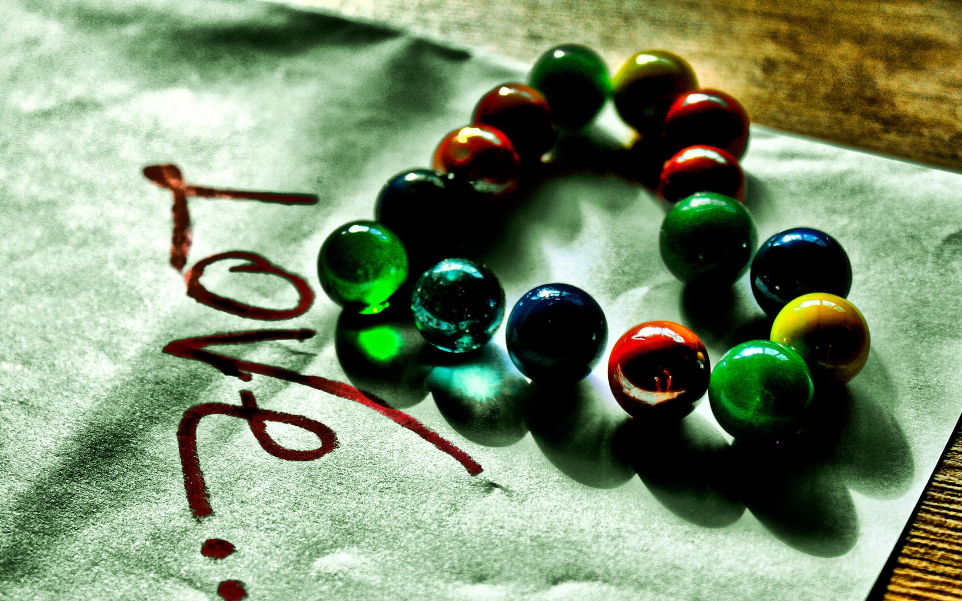 An artistic HD wallpaper featuring colorful marbles arranged in a heart shape on paper, with the word Love written boldly beneath them.