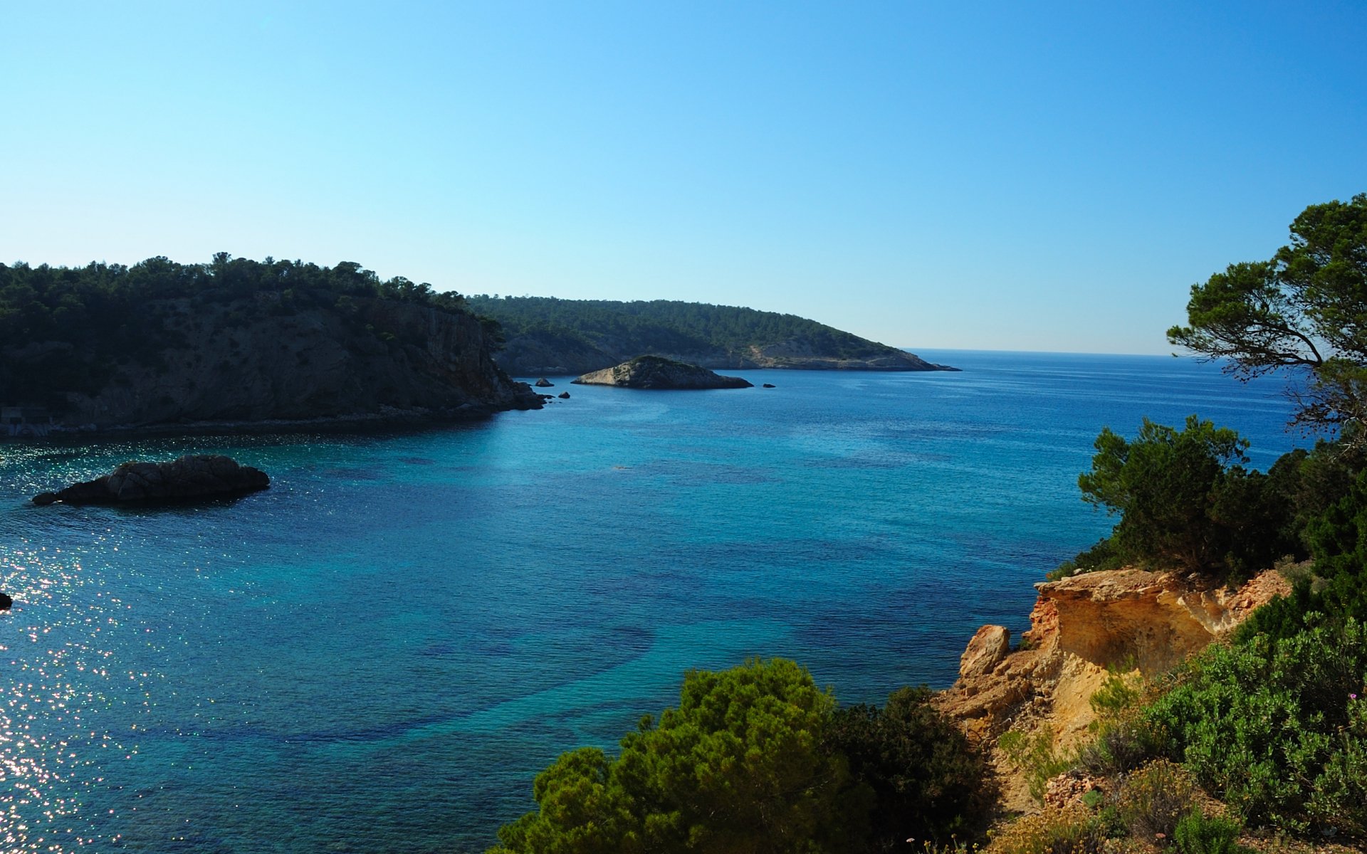 HD PC desktop wallpaper showcasing a serene nature seascape with clear blue waters, rocky coastline, and lush greenery under a bright, cloudless sky.
