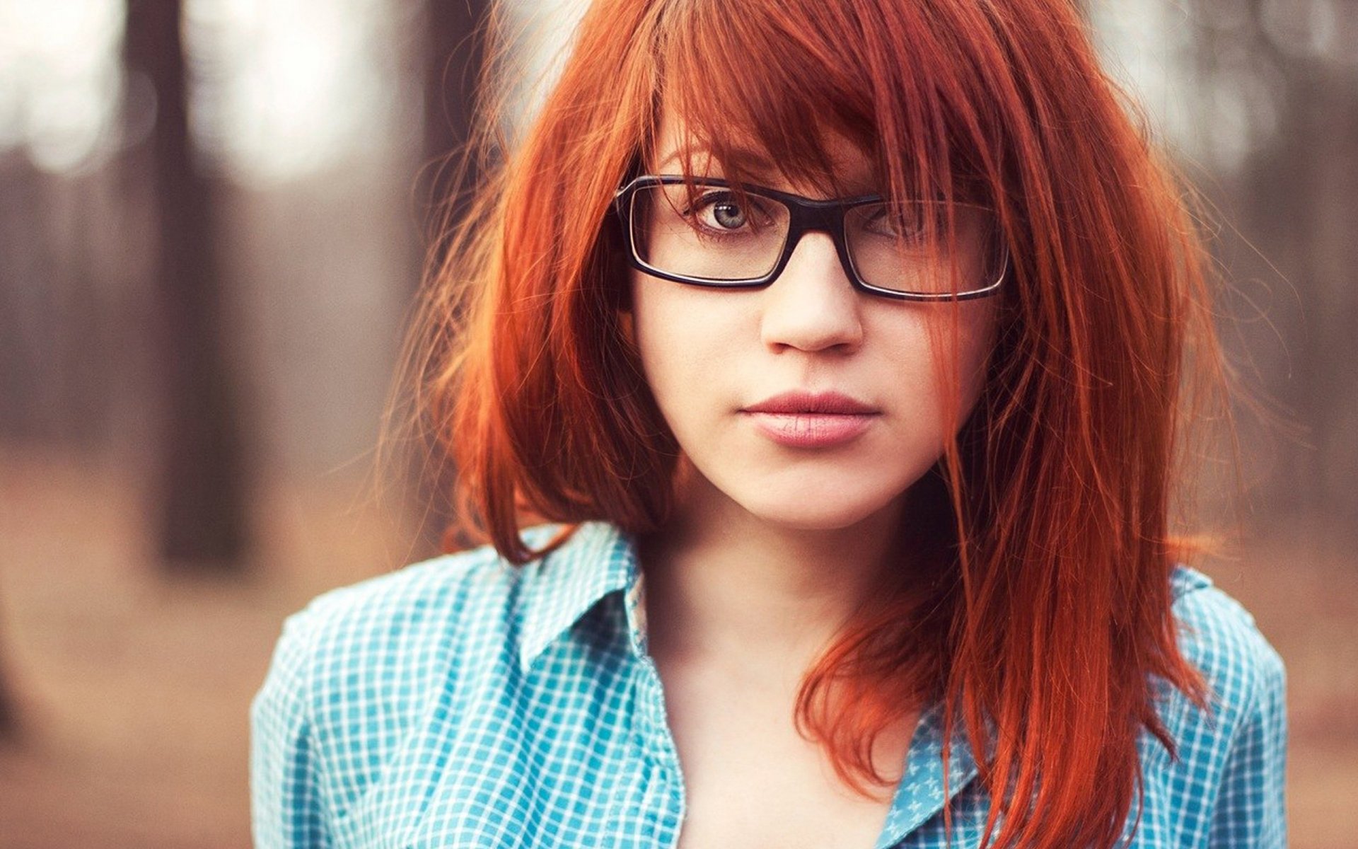 A beautiful redhead woman with glasses gazes confidently at the camera, surrounded by a serene forest backdrop, creating an enchanting HD desktop wallpaper.
