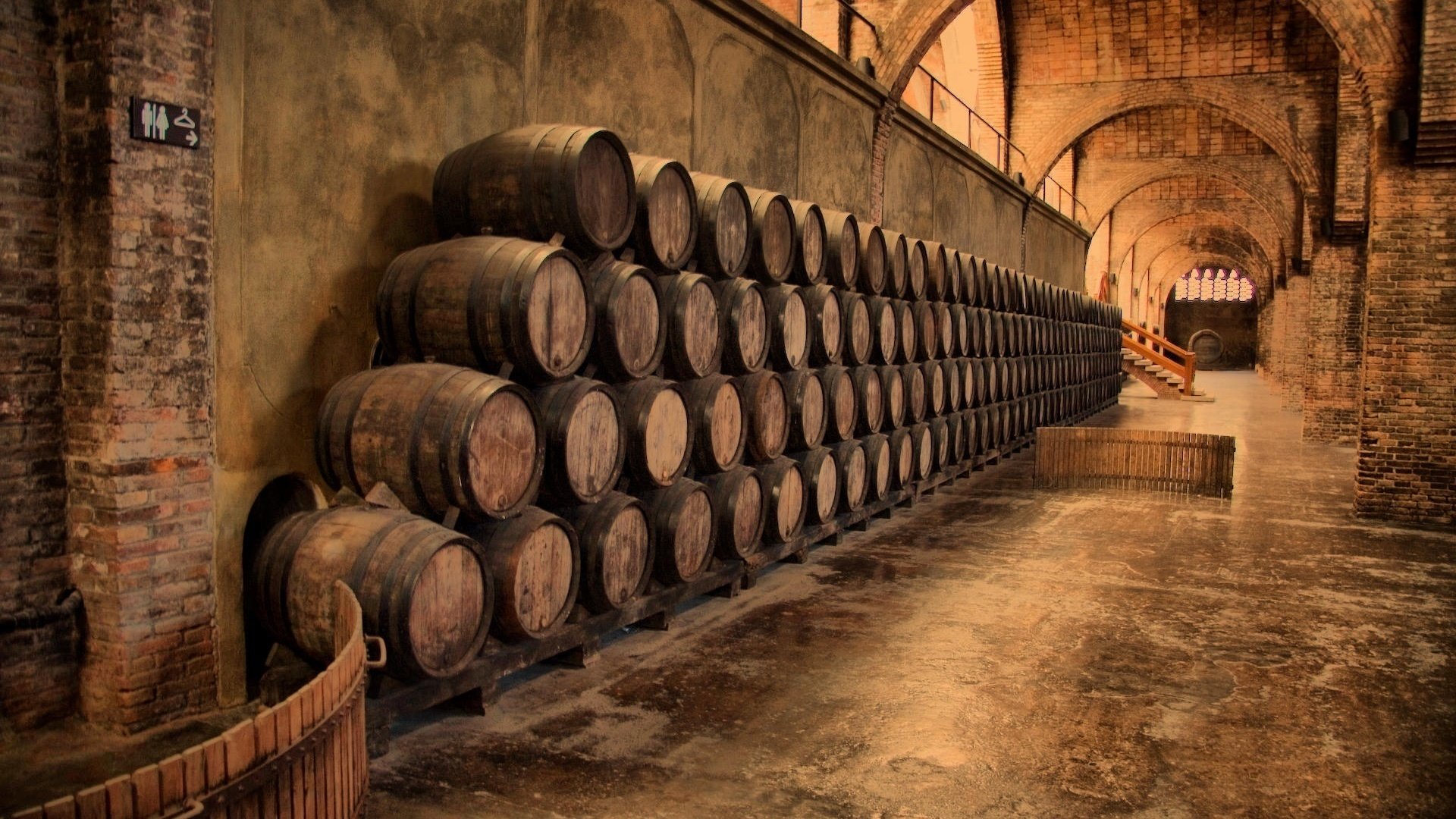 A serene HD wallpaper showcasing a rustic wine cellar with neatly stacked wooden barrels against brick walls, highlighting the craftsmanship of man-made structures in a historical place.
