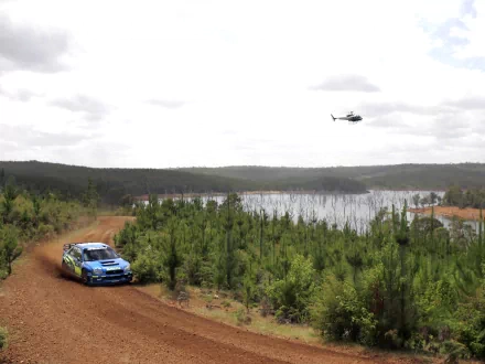 Rally race car speeds on a dirt road near Lake Bled, surrounded by forest, with a helicopter hovering above, captured in an HD desktop wallpaper.