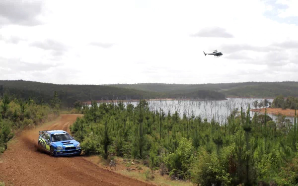 Rally race car speeds on a dirt road near Lake Bled, surrounded by forest, with a helicopter hovering above, captured in an HD desktop wallpaper.
