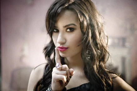 HD desktop wallpaper featuring singer Demi Lovato with brunette hair, brown eyes, and a shushing gesture against a soft, blurred background.