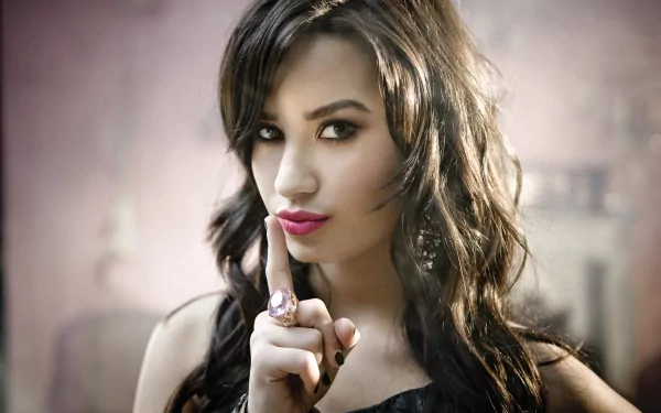 HD desktop wallpaper featuring singer Demi Lovato with brunette hair, brown eyes, and a shushing gesture against a soft, blurred background.
