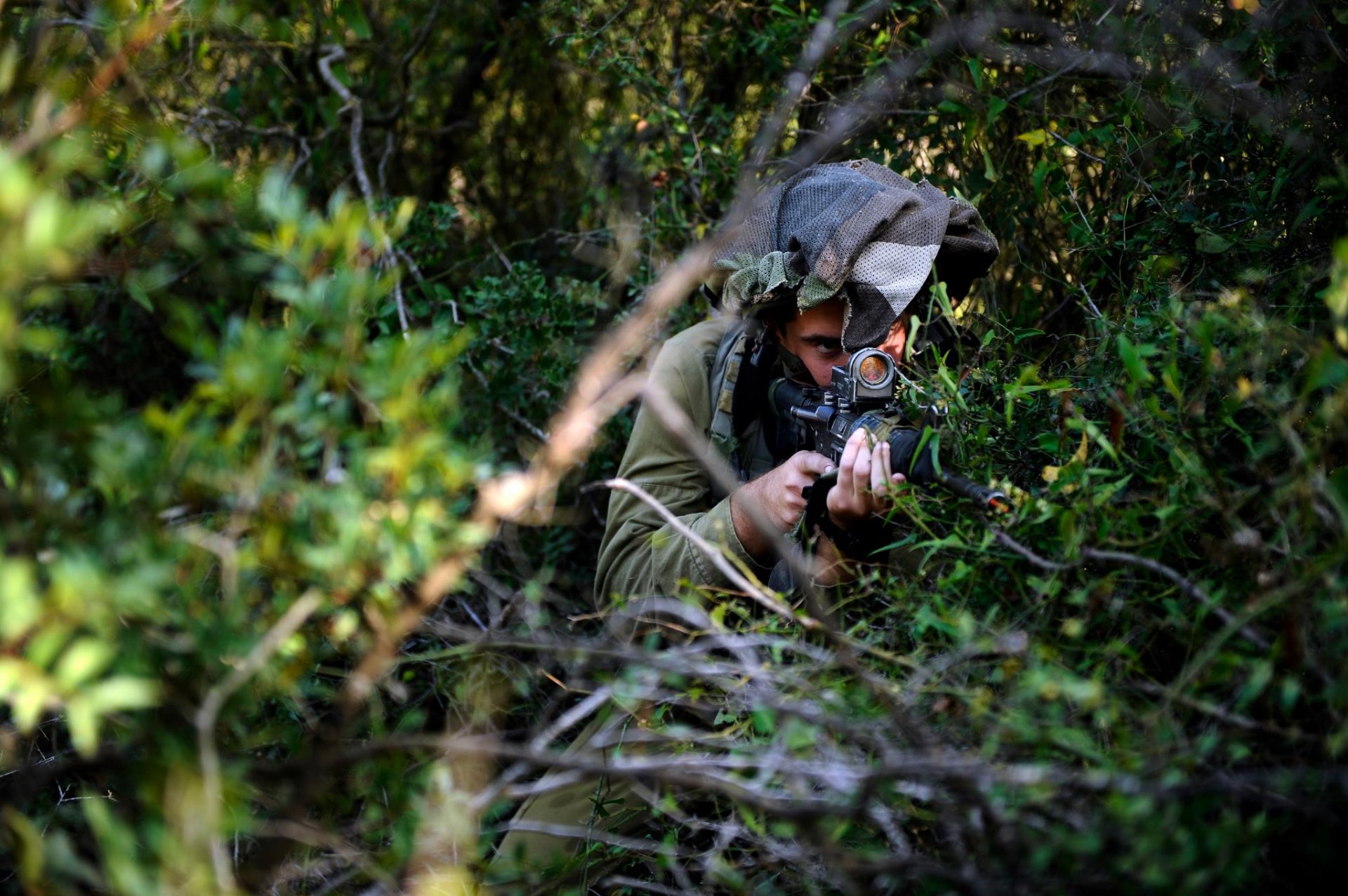 HD PC desktop wallpaper/background: military soldier in camouflage and ghillie hood aiming a rifle while concealed in dense green foliage.